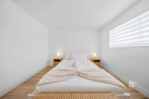 Une chambre d’hôtel au style minimaliste, dotée d’un grand lit, de deux tables de chevet avec des lampes et d’une fenêtre avec des stores.