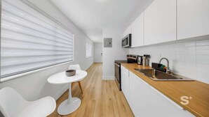 Een moderne keuken met een houten aanrecht, witte kasten, een ingebouwde magnetron en een klein zithoekje met een witte tafel en stoelen.