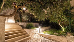 Un escalier en pierre mène à un jardin sur le toit, agrémenté de murs en pierre, de plantes en pot et d’un arbre aux fleurs roses.