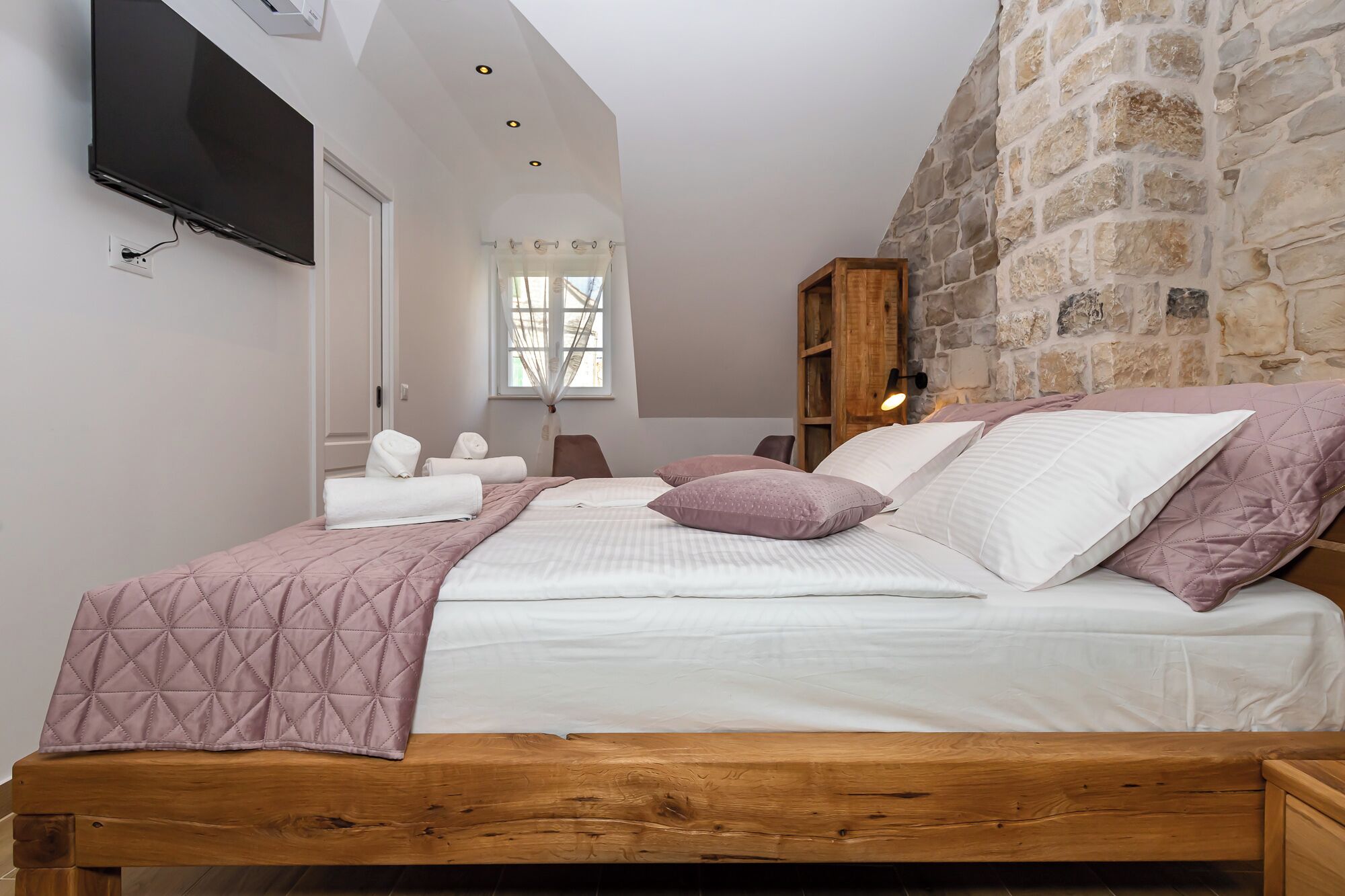 A bedroom with a wooden bed, white bedding, and a stone wall.