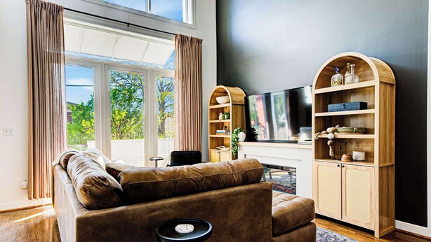 Ein Wohnzimmer mit einer Ledersofa, einem Fernseher, Holzregalen und einem großen Fenster mit Vorhängen.