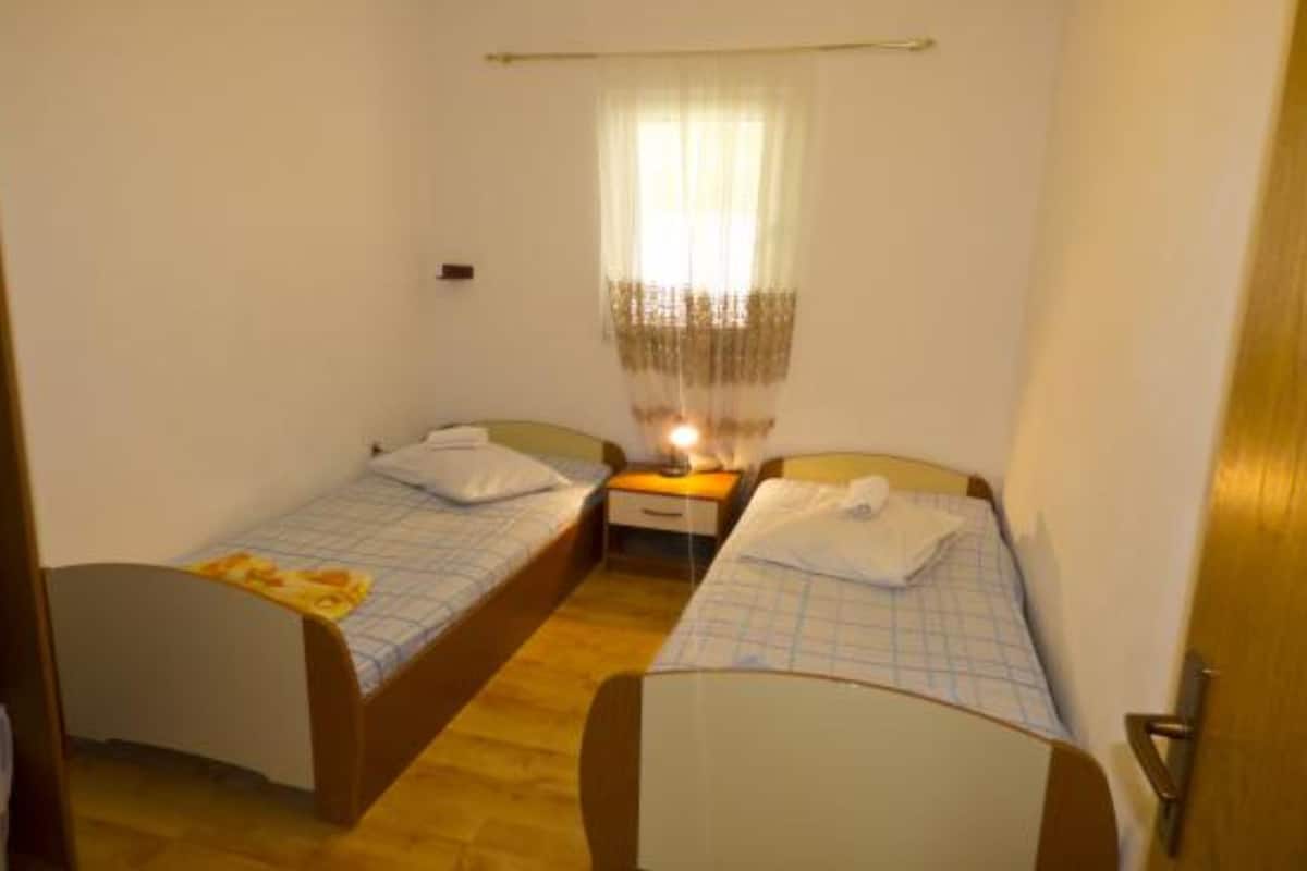 Une petite chambre d’hôtel composée d’une seule pièce, avec deux lits simples, une table de chevet avec une lampe et une fenêtre ornée de rideaux légers.