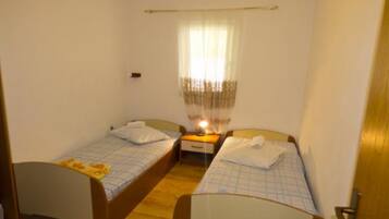 A small, single-room hotel room with two single beds, a bedside table with a lamp, and a window with sheer curtains.