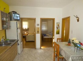 Cocina compacta con refrigerador, lavamanos y televisor. Se observa un comedor con mesa y sillas, y una habitación visible a través de una puerta abierta.