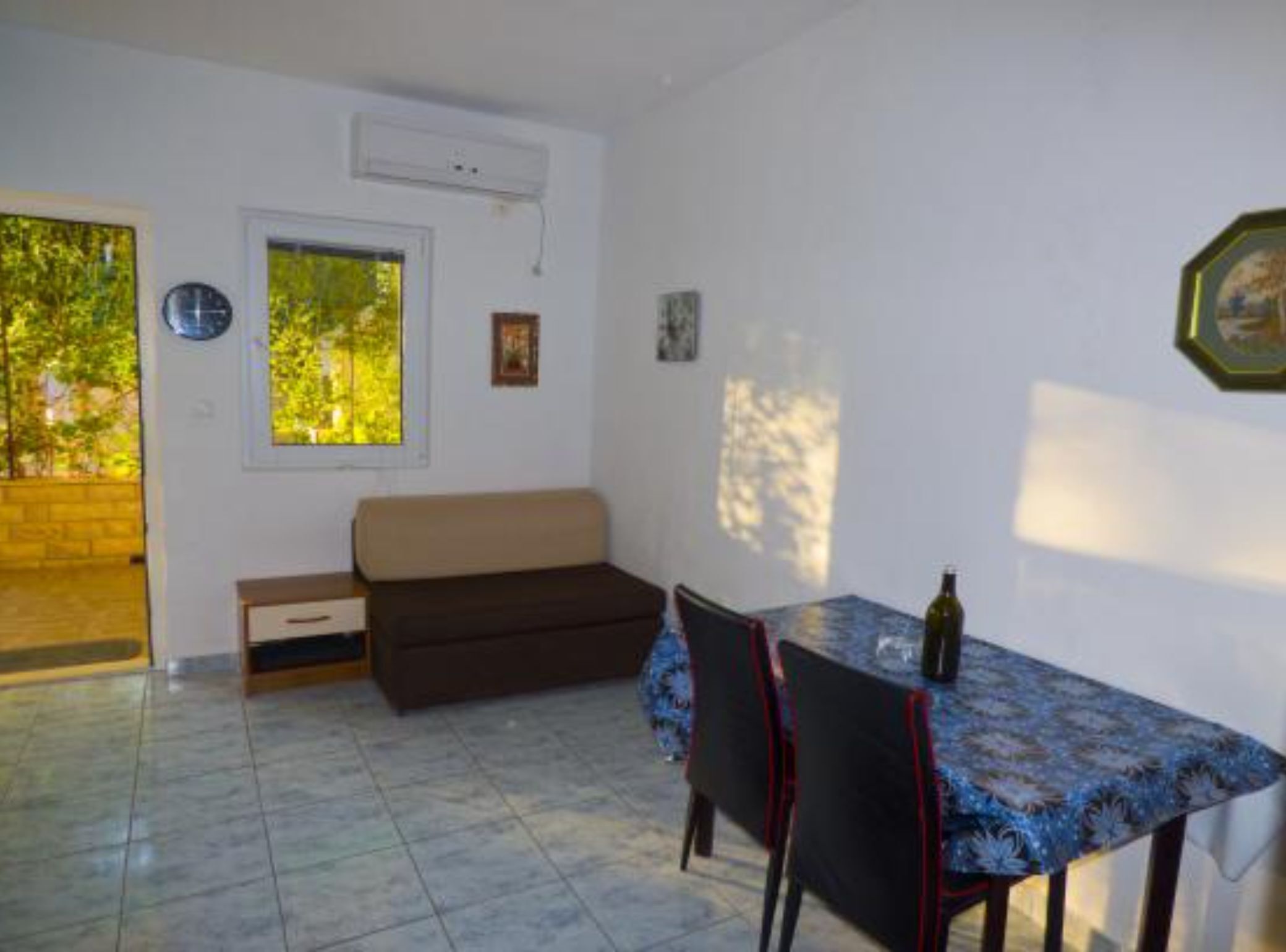 A small, bright room with a tiled floor, a sofa, a dining table with chairs, and a window with a view of greenery.