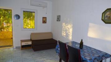 A small, bright room with a tiled floor, a sofa, a dining table with chairs, and a window with a view of greenery.
