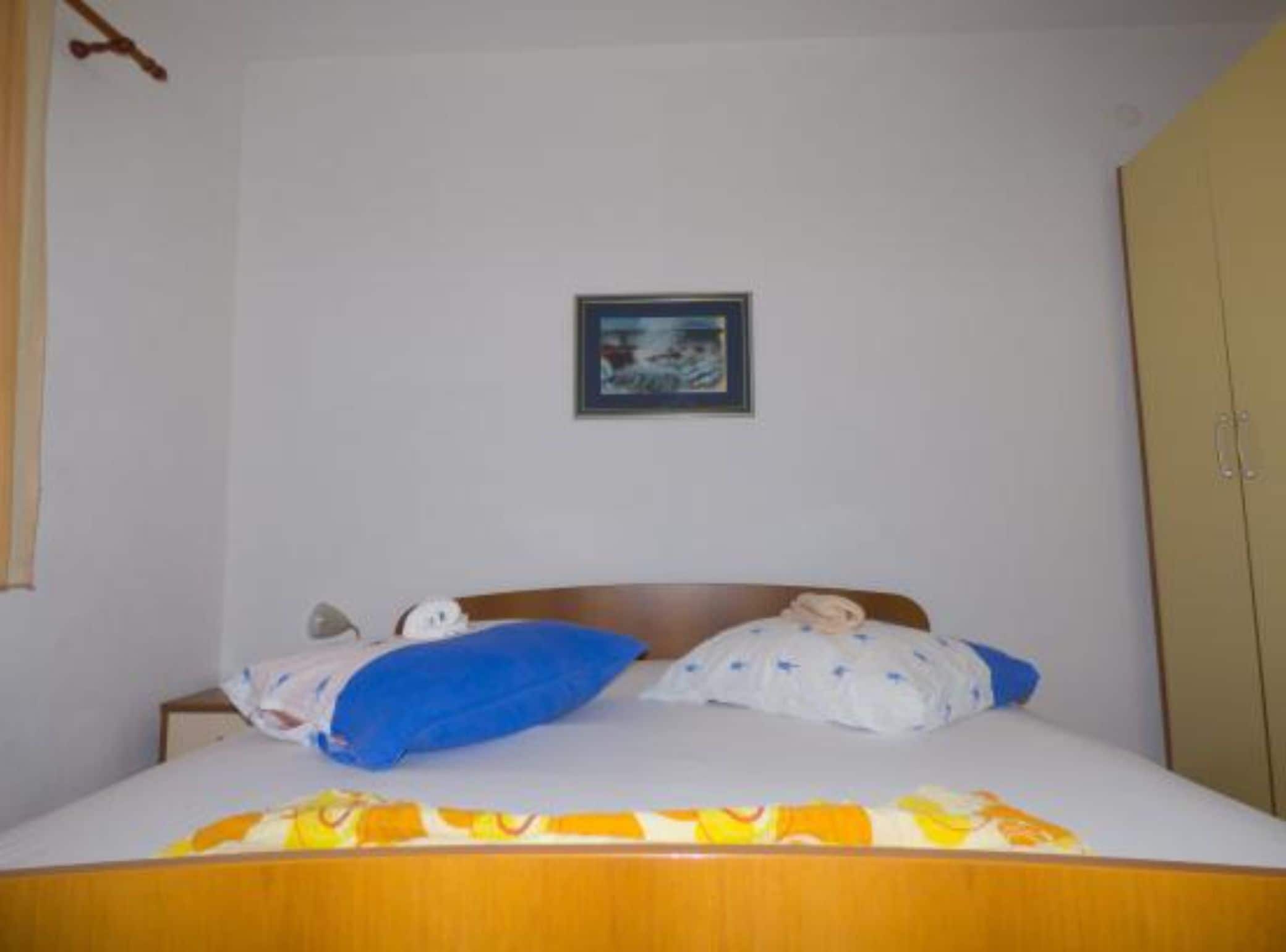 Una cama individual con cabecera de madera, colchón blanco y almohada azul. Encima de la cama, hay un cuadro enmarcado en la pared.
