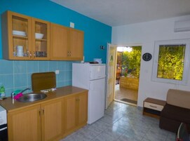 Cocina compacta con gabinetes de madera, un refrigerador y un lavamanos. Cuenta con una pequeña zona de comedor con una mesa y sillas. Una ventana ofrece vistas al exterior.