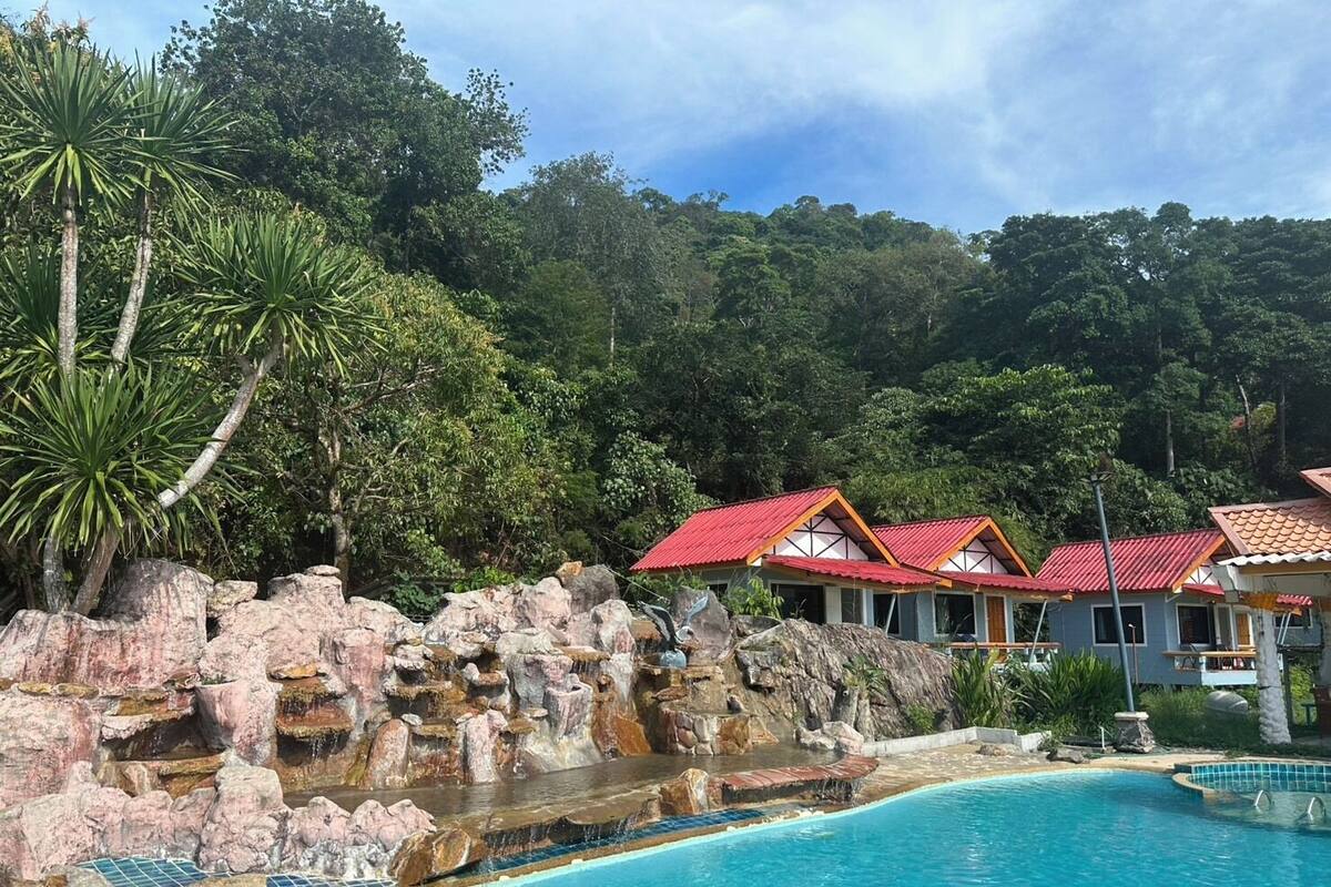 Une piscine agrémentée d’une cascade artificielle entourée de rochers, le tout niché au milieu d’une végétation luxuriante et bordé de bâtiments aux toits rouges.