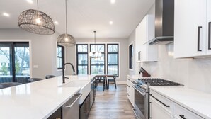 A modern kitchen with white cabinetry, a large island, stainless steel appliances, and pendant lighting. - Serenity Now: Grand Beach Retreat, Heated Spa Pool, Hot Tub, Sauna, Golf Cart (New Buffalo)