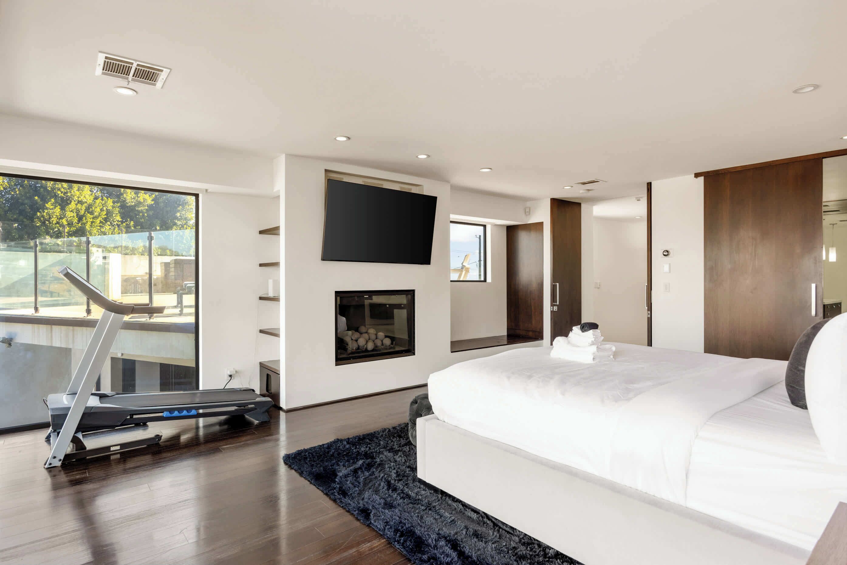 A modern bedroom with a large bed, a treadmill, a fireplace, and a TV mounted on the wall.