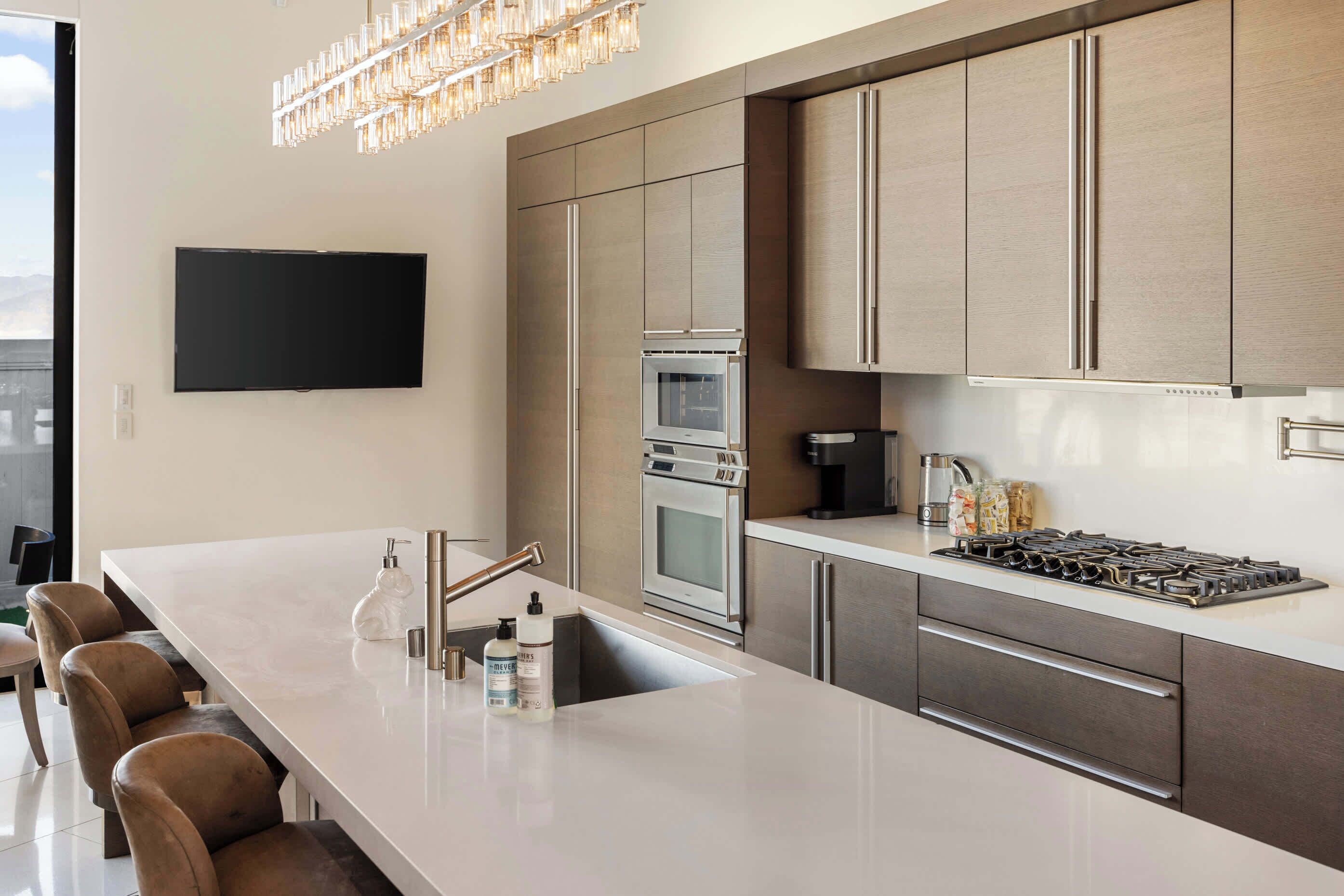 A modern kitchen with a large island, stainless steel appliances, and a flat-screen TV mounted on the wall.