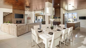 A modern kitchen with a central island, a dining area with a white table and chairs, and a living area with a sofa and TV.