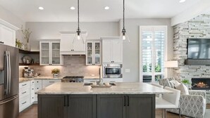 A modern kitchen with a large island, white cabinetry, and a stone fireplace.