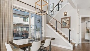 A modern dining area with a wooden table and white chairs, a staircase with a wooden railing, and a view of the outside through large windows.
