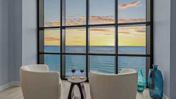 Een kamer met een groot raam met uitzicht op zee, twee stoelen, een klein tafeltje met een wijnglas, en twee decoratieve vazen.