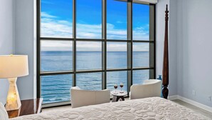 Een slaapkamer met een groot raam met uitzicht op zee, een bed met een geborduurd dekbed, twee stoelen en een klein rond tafeltje met glazen.