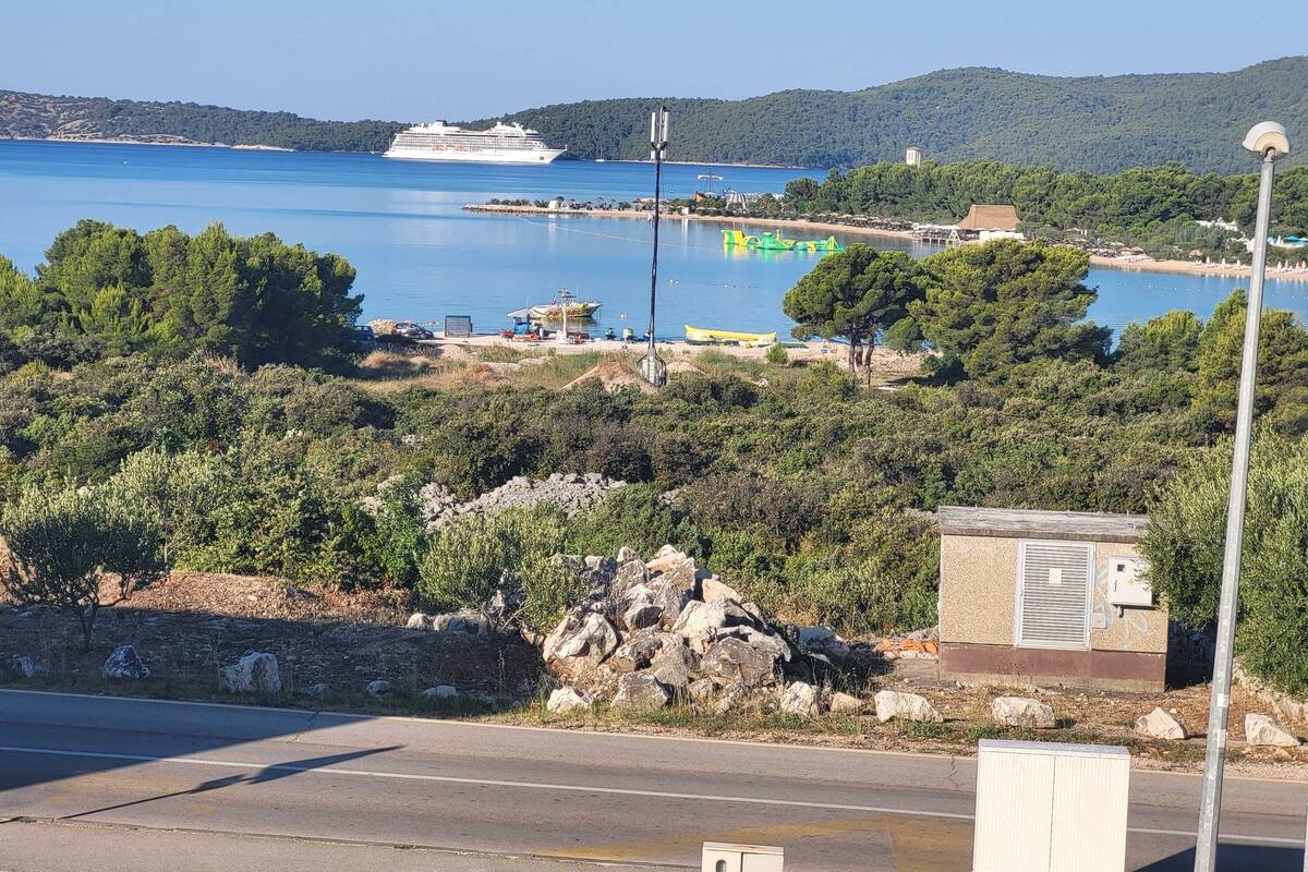 Eine Küstenstraße mit freiem Blick auf ein großes Kreuzfahrtschiff im Hintergrund, ein kleiner Hafen mit Booten und eine felsige Küstenlinie mit spärlicher Vegetation.