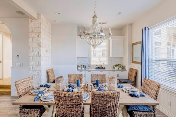 A dining area with a large table set for six, surrounded by wicker chairs, a chandelier, and a view of the kitchen in the background.