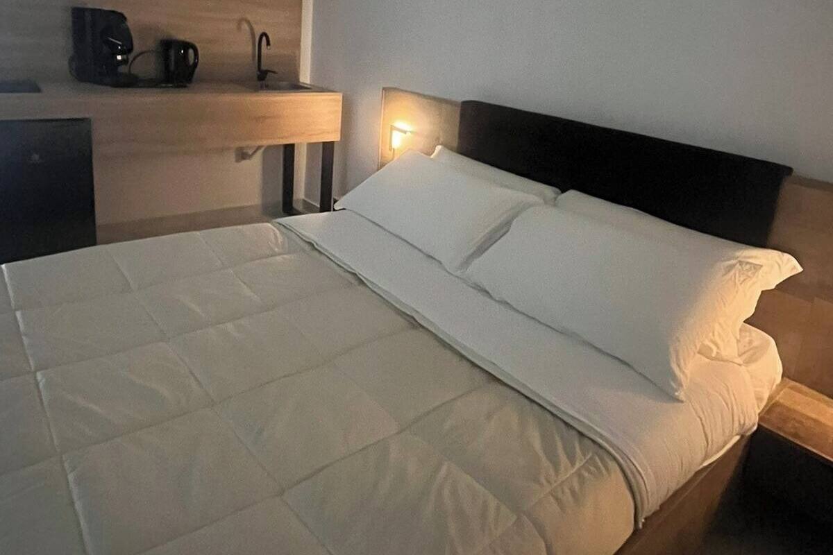A neatly made bed with white bedding and a headboard, a bedside table with a lamp, and a coffee maker on a shelf.