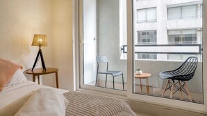 Una camera d'albergo con un letto, una lampada, una sedia e un balcone con vista su un edificio.