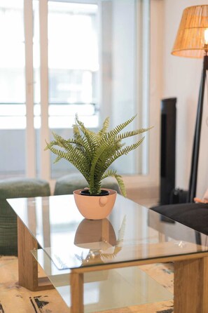 Een glazen tafel met een varenpot in het midden, een lamp aan de rechterkant en een groene bank op de achtergrond.