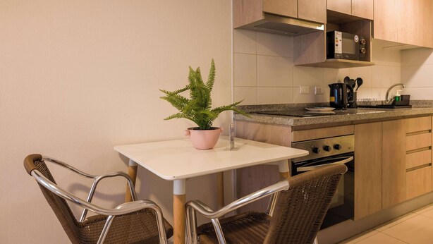 Cocina moderna con una mesa blanca, dos sillas de mimbre, un helecho en maceta y electrodomésticos de acero inoxidable.