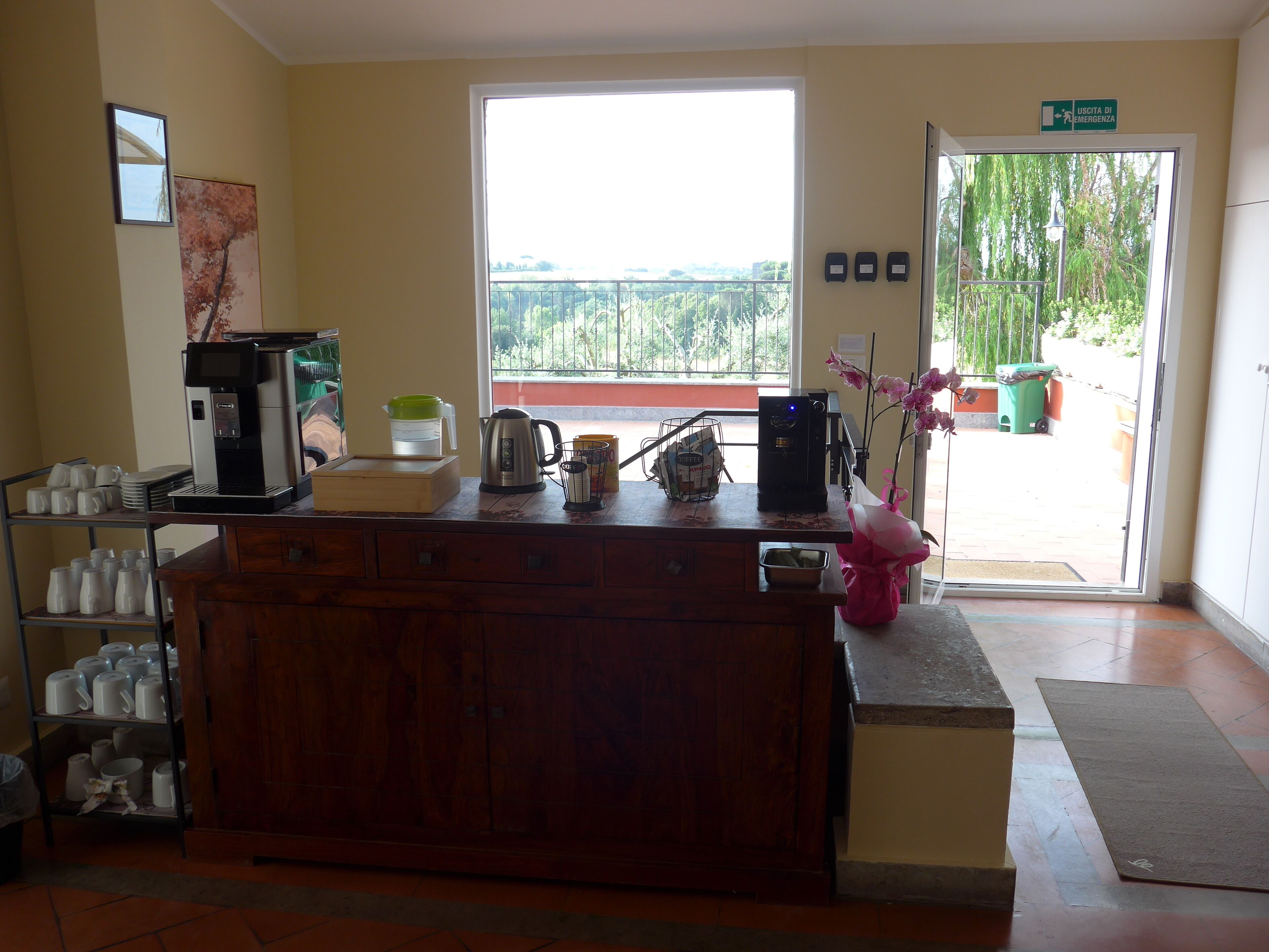 Zona de recepción con máquina de café, hervidor y un jarrón de flores. Se observa una ventana con vistas a zonas verdes y una puerta con la leyenda 'USCITA DI EMERGENZA'.