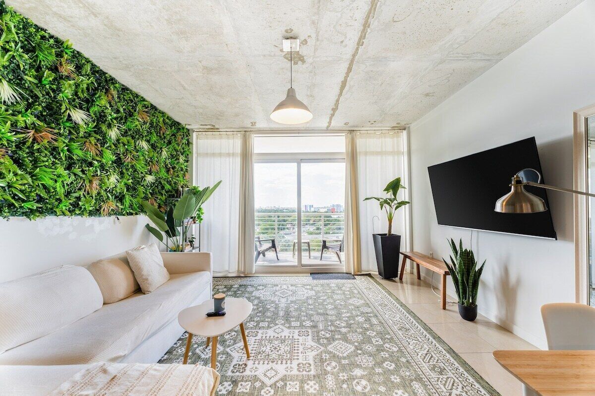 Een moderne woonkamer met een witte bank, een salontafel, een kamerkleed met dessin, een grote televisie en een groene wand met planten.
