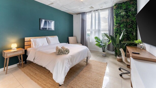 A modern bedroom with a bed, bedside table, lamp, and a large window with curtains. - 1BR Miami Retreat w/BBQ, Pool, & Skyline Views (Miami)