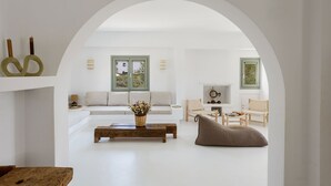 Amplia sala de estar con un sofá blanco, una mesa de centro de madera y un pouf tejido. Hay dos ventanas con marcos verdes y una lámpara fijada a la pared.
