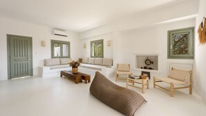 Amplia sala de estar con un sofá blanco, una mesa de centro de madera y un taburete tejido. Se observan dos ventanas con marcos verdes, una chimenea y un aire acondicionado de pared.