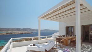 Un balcón con mobiliario blanco, una mesa de madera y vistas al mar y a las montañas.