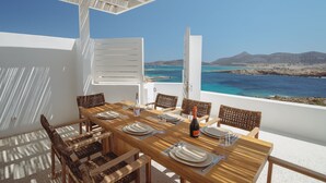 A dining table set for four with wicker chairs, a bottle of wine, and a view of the sea and mountains.