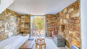 A room with stone walls, a wooden table, two chairs, a bench, and a large window with a view of greenery. - Stone Pine House | Antiparos (Soros)