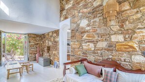 A room with a stone wall, a wooden sofa with cushions, a small table, and a chair. - Stone Pine House | Antiparos (Soros)