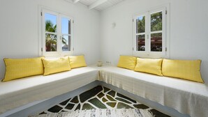 A room with two white sofas with yellow patterned cushions, a white ceiling with wooden beams, and a view of palm trees and a mountain through the windows. - Orchard House | Antiparos (Agios Georgios)