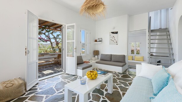 A spacious living area with a stone floor, a grey sofa, a white coffee table, and a view of the outdoors through an open door. - Orchard House | Antiparos (Agios Georgios)
