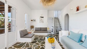 A modern living room with a stone-patterned floor, a grey sofa, a white sofa with blue pillows, and a hanging light fixture. - Orchard House | Antiparos (Agios Georgios)