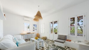 A bright, airy room with white walls and a stone-patterned floor. There is a white sofa, a small table with a bowl of fruit, and a hanging light fixture. - Orchard House | Antiparos (Agios Georgios)