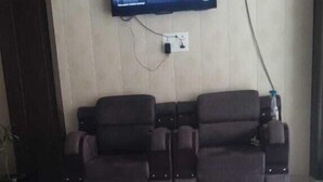 Een kamer met een wandgemonteerde flatscreen-tv, een donkerbruine bank en een betegelde vloer.