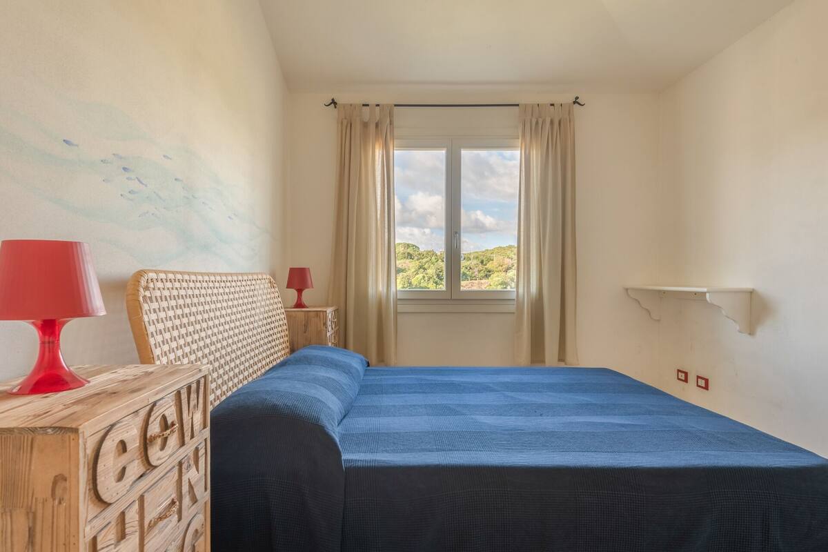 A bedroom with a bed, a wooden dresser, a red lamp, a window with curtains, and a view of greenery outside.