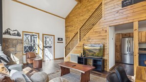 A modern living room with a wooden staircase, a stone wall, and a kitchen area visible through an open door. - High Top Cabin with King Beds, Fire Pits, Hot Tub, Arcade, 🐻 Kids’ Clubhouse (Sevierville)