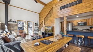 A cozy interior with a wooden staircase, a stone fireplace, a dining area with a wooden table, and a kitchen with wooden cabinets and a bar. - High Top Cabin with King Beds, Fire Pits, Hot Tub, Arcade, 🐻 Kids’ Clubhouse (Sevierville)
