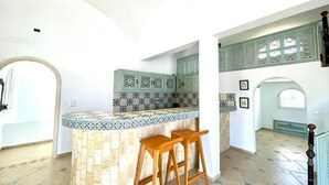 A kitchen with a central island, two wooden bar stools, patterned backsplash, and light green cabinets. - Villa Dar Othman (cap zebib)