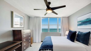 A hotel room with a bed, a desk, a dresser, a ceiling fan, and a large window with a view of the ocean.