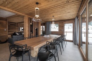 Ein großzügiger Essbereich mit einem langen Holztisch, modernen Stühlen, großen Fenstern und einer Holzbalkendecke.