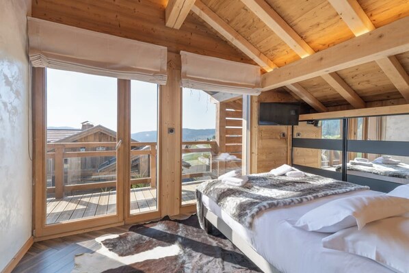 A bedroom with a large bed, wooden ceiling beams, and a view of a wooden deck and buildings.
