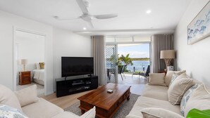 A modern living room with a flat-screen TV, a wooden coffee table, and a sofa. There is a balcony with a view of the water.