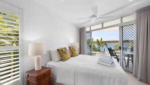 A bedroom with a large bed, two nightstands, a ceiling fan, and a view of a body of water through large windows.